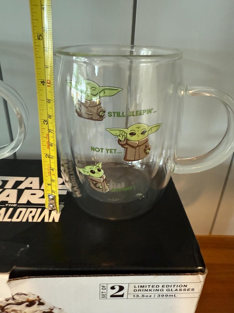 K&ouml;nnte beinhalten: Klarer Glasbecher mit Grogu aus The Mandalorian, mit den Aufschriften "STILL SLEEPIN'" und "NOT YET...". Der Becher ist etwa 10 cm hoch und hat einen Henkel. Der Becher ist Teil eines Sets von zwei Trinkgl&auml;sern in limitierter Auflage.