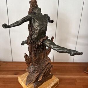 May include: Bronze sculpture of a figure with outstretched arms and legs, emerging from a textured, tree-like form. The sculpture is mounted on a rectangular, light-colored stone base. The figure has a dark, aged patina.