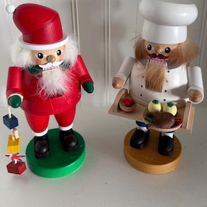May include: Two wooden figurines: Santa Claus and a chef. Santa wears a red suit, hat, and has a white beard. The chef wears a white uniform and hat, holding a tray with food. Both figures have black shoes and are on round bases.