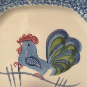 May include: A blue rooster with a red comb and green tail feathers is painted on a white ceramic plate. The rooster is standing on a green branch with a blue pond below.