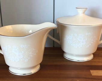 May include: A cream-colored creamer and sugar bowl set with a delicate floral design. The set is made of fine china and has a gold trim.