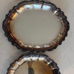May include: Three silver-toned metal serving trays with ornate, embossed floral borders. The collection includes one rectangular tray with two side handles and two oval-shaped trays of varying sizes, all featuring a reflective, tarnished finish.