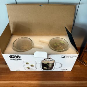 K&ouml;nnte beinhalten: Ein Karton mit zwei transparenten Glasbechern mit Griffen. Die Becher zeigen ein Design von Grogu aus The Mandalorian. Die Box hat das Star Wars Mandalorian Collection Logo und den Text "Limited Edition Drinking Glasses".