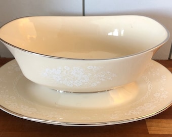 May include: A white porcelain gravy boat with a silver rim and a floral design. The boat is sitting on a matching saucer.