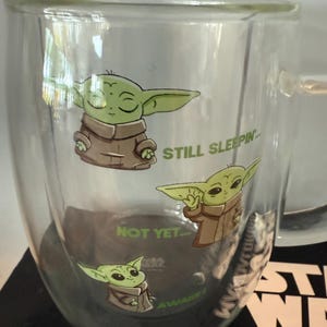 K&ouml;nnte beinhalten: Doppelwandige Glas-Tasse mit Illustrationen eines gr&uuml;nen Alien-Charakters in einem braunen Gewand. Auf der Tasse stehen die S&auml;tze "STILL SLEEPIN'", "NOT YET..." und "AWAKE!". Die Tasse hat einen Henkel und ist wahrscheinlich f&uuml;r Hei&szlig;getr&auml;nke.