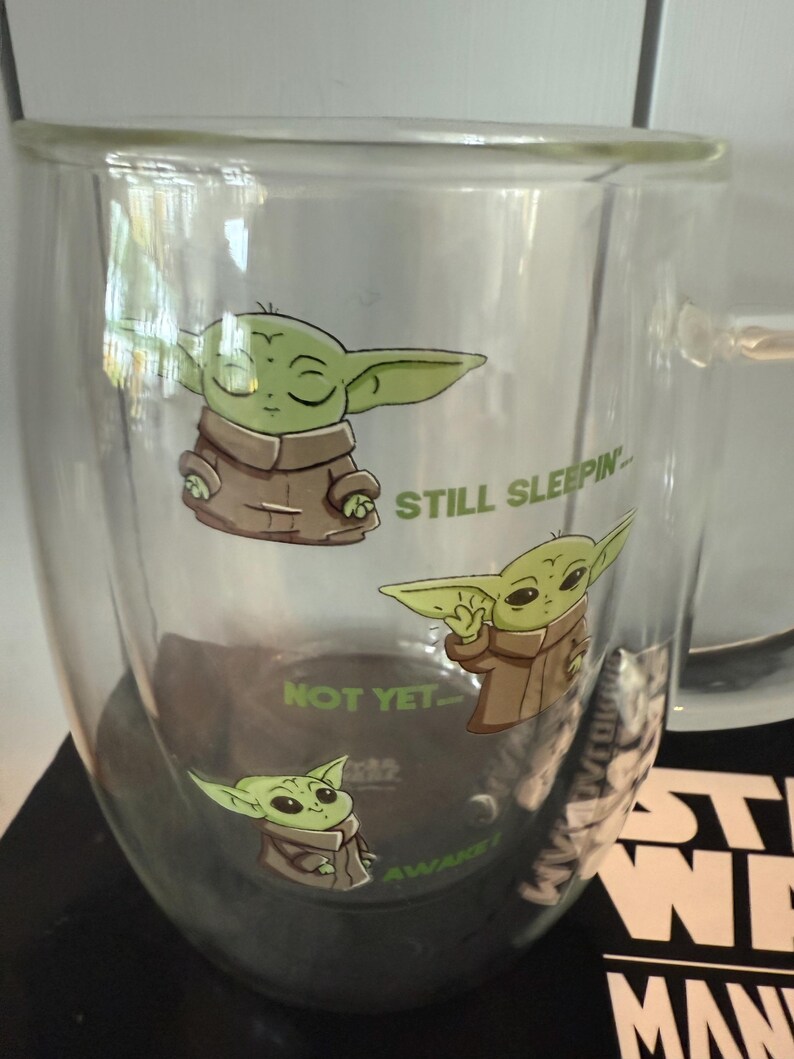 K&ouml;nnte beinhalten: Doppelwandige Glas-Tasse mit Illustrationen eines gr&uuml;nen Alien-Charakters in einem braunen Gewand. Auf der Tasse stehen die S&auml;tze "STILL SLEEPIN'", "NOT YET..." und "AWAKE!". Die Tasse hat einen Henkel und ist wahrscheinlich f&uuml;r Hei&szlig;getr&auml;nke.