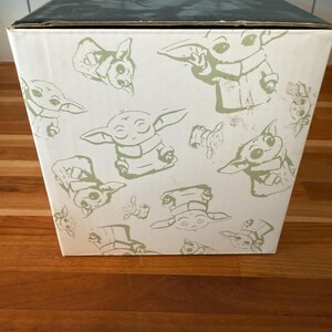 K&ouml;nnte beinhalten: Eine wei&szlig;e Box mit schwarzem Deckel mit einem sich wiederholenden Muster aus gr&uuml;n umrandeten Illustrationen einer Figur aus einem beliebten Science-Fiction-Franchise. Die Box steht auf einer Holzoberfl&auml;che.
