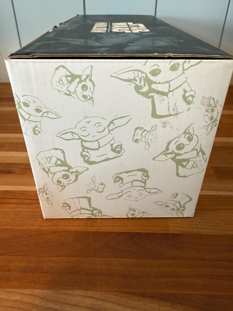 K&ouml;nnte beinhalten: Eine wei&szlig;e Box mit schwarzem Deckel mit einem sich wiederholenden Muster aus gr&uuml;n umrandeten Illustrationen einer Figur aus einem beliebten Science-Fiction-Franchise. Die Box steht auf einer Holzoberfl&auml;che.