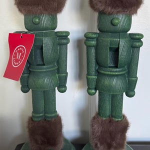 May include: Two green wooden nutcracker figures with textured wood grain finishes. Each wears a tall brown faux fur hat and matching brown faux fur leg cuffs. A red tag with the white text Martha Stewart is attached to the left nutcracker.