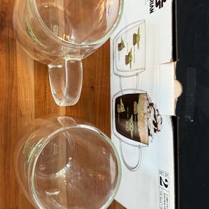 K&ouml;nnte beinhalten: Zwei transparente Glasbecher mit Griffen und einem gr&uuml;nen Charakterdesign stehen auf einer Holzoberfl&auml;che. Eine Schachtel mit dem Branding "Star Wars Mandalorian" ist teilweise sichtbar, was auf ein Produkt mit Thema hindeutet. Die Becher geh&ouml;ren zu einem Set.