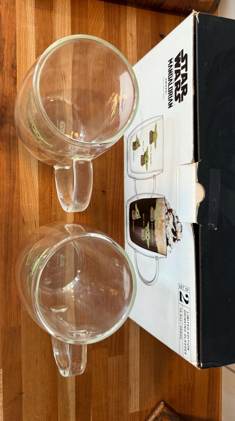 K&ouml;nnte beinhalten: Zwei transparente Glasbecher mit Griffen und einem gr&uuml;nen Charakterdesign stehen auf einer Holzoberfl&auml;che. Eine Schachtel mit dem Branding "Star Wars Mandalorian" ist teilweise sichtbar, was auf ein Produkt mit Thema hindeutet. Die Becher geh&ouml;ren zu einem Set.