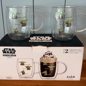 K&ouml;nnte beinhalten: Zwei transparente Glasbecher mit Griffen, die Illustrationen von Grogu aus The Mandalorian zeigen. Jeder Becher hat drei Grogu-Bilder und die S&auml;tze "Still Sleeping" und "Not Yet". Die Becher stehen auf einer Star Wars Mandalorian-Box.