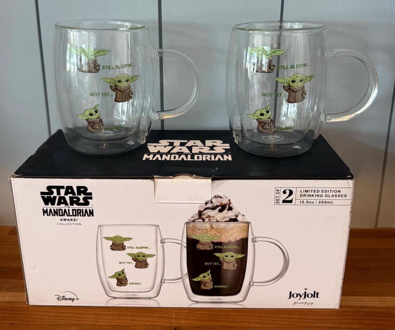 K&ouml;nnte beinhalten: Zwei transparente Glasbecher mit Griffen, die Illustrationen von Grogu aus The Mandalorian zeigen. Jeder Becher hat drei Grogu-Bilder und die S&auml;tze "Still Sleeping" und "Not Yet". Die Becher stehen auf einer Star Wars Mandalorian-Box.