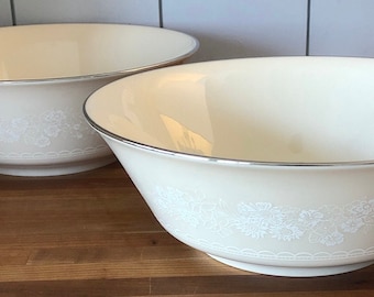 May include: Two white ceramic bowls with a silver rim and a floral pattern. The bowls are on a wooden surface.