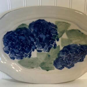 May include: An oval-shaped ceramic serving platter with a glossy off-white finish. The center features a hand-painted design of three clusters of deep blue hydrangea flowers surrounded by textured green leaves, with a scalloped edge detail.