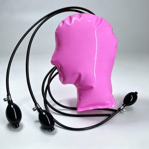 Peut inclure: Un masque gonflable rose vif en forme de tête avec une finition lisse et brillante. Trois fins tubes noirs partent du sommet de la tête, chacun relié à une petite pompe manuelle noire de forme ovale avec une valve métallique de contrôle.