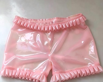 Könnte beinhalten: Hellrosa, durchscheinende Shorts mit glänzender Oberfläche und Rüschenbesatz am Bund sowie an den Beinabschlüssen. Das Kleidungsstück hat eine glatte, glänzende Textur und eine durchgehend einheitliche, blassrosa Farbe.