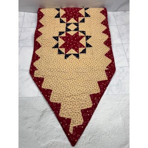 May include: A quilted table runner featuring a cream-colored fabric with a small dark floral print, accented by deep red fabric with a white star and crescent moon pattern. The design includes dark navy blue star-shaped patchwork elements.