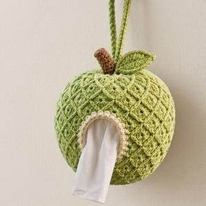 May include: A round, light green crocheted apple-shaped tissue dispenser featuring a textured diamond pattern, a brown stem, and a single green leaf. A white tissue is pulled through the circular opening, which is trimmed with a cream-colored border.
