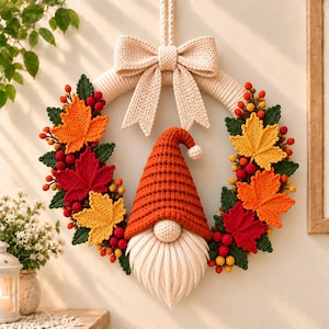 Può includere: Una ghirlanda circolare con un fiocco beige all'uncinetto sulla parte superiore, decorata con foglie autunnali all'uncinetto nei colori arancione, rosso e giallo, insieme a piccole bacche rosse e gialle. Un gnomo centrale all'uncinetto indossa un alto cappello arancione.
