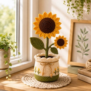 May include: A crocheted sunflower plant with a large and a small flower, featuring yellow petals and dark brown centres. The plant sits in a cream-coloured crocheted pot with a scalloped rim, a brown drawstring, and small embroidered yellow flowers.