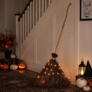May include: A tall decorative broom made of natural brown twigs with a long wooden handle, illuminated by small warm white fairy lights woven throughout the bristles. The broom stands upright, creating a rustic display piece for seasonal decor.
