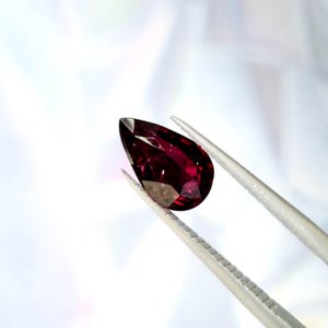 Puede incluir: Una piedra preciosa de color rojo intenso en forma de pera sujeta por unas pinzas de metal plateado. La piedra facetada muestra un tono carmesí oscuro con reflejos de luz internos y bordes afilados, con una superficie clara y pulida.