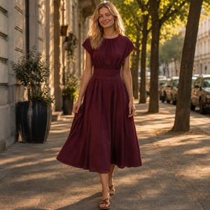 Può includere: Un abito midi color bordeaux intenso con scollo rotondo, maniche corte e vita aderente con una fascia larga. La gonna è plissettata e presenta una silhouette svasata che arriva sotto le ginocchia, abbinato a sandali marroni.
