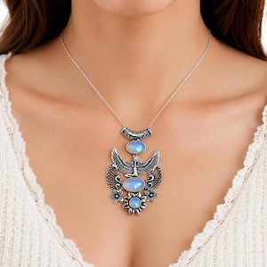 May include: A silver-toned necklace featuring a large pendant with three oval iridescent moonstones. The pendant design includes a central winged figure, intricate filigree patterns, and small floral accents at the base, suspended from a thin silver chain.
