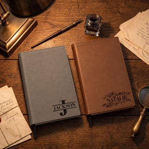 May include: Two rectangular journals with smooth covers, one in grey and one in brown. The grey journal features the name JACKSON with a large letter J. The brown journal features the name NATALIE surrounded by a decorative leaf and vine illustration.