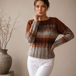 Puede incluir: Un suéter de punto de manga larga con textura acanalada y rayas horizontales de bloques de color en tonos marrón óxido, marrón oscuro y gris claro. La prenda tiene un cuello redondo y una silueta ajustada, combinada con pantalones blancos lisos.