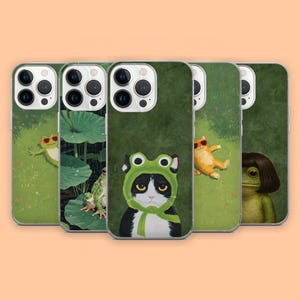 May include: A collection of five smartphone cases featuring various illustrations: a frog wearing sunglasses, frogs on lily pads, a black and white cat wearing a green frog hood, an orange cat wearing sunglasses, and a frog with a brown bob haircut.
