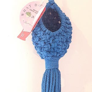 May include: A blue macrame hanging basket with a knotted texture and a long tassel at the bottom. The basket is suspended by a matching blue cord and features a round opening. A pink circular tag is attached, displaying the text WHAT DO.