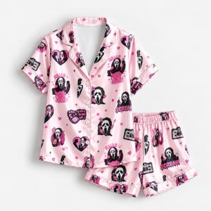 Könnte beinhalten: Ein hellrosa Pyjama-Set bestehend aus einem kurzärmeligen Hemd mit Knopfleiste und passender Shorts. Der Stoff ist mit einem wiederkehrenden Muster aus der Ghostface-Maske, Messern, Herzen sowie dem Text "HELLO SIDNEY" und "DO YOU HANG UP" bedruckt.