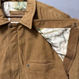 May include: A tan suede jacket featuring a pointed collar, a front button closure, and a welt pocket. The interior lining is cream-colored fabric printed with a detailed map design, including a compass rose and text labels like Last Chance Gap.