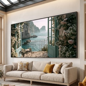 May include: A large rectangular wall art print featuring an open teal window frame looking out onto a coastal Mediterranean scene with rocky cliffs, blue sea, and a balcony with a small black metal table, surrounded by climbing white and pink roses.