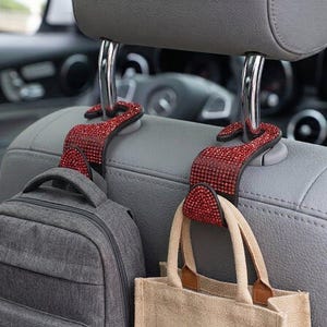 Op de afbeelding: Twee S-vormige haken voor autostoelsteunen, bedekt met kleine, fonkelende rode strass-steentjes. Elke haak is bevestigd aan de metalen stangen van een grijze hoofdsteun. Een grijze rugzak hangt aan de linkerhaak en een beige tas aan de rechterhaak.