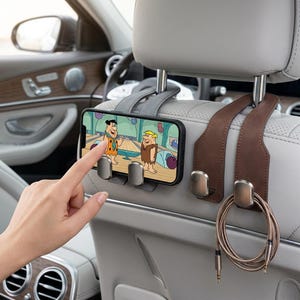 May include: A gray and brown leather car headrest hook set with integrated phone mount. The mount holds a smartphone displaying a cartoon scene with two characters. A coiled charging cable hangs from one of the metallic hooks on the brown strap.
