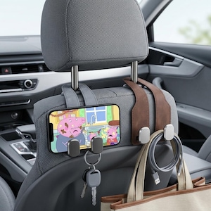 May include: A car headrest organiser set featuring two leather-like straps with metal hooks. One strap holds a smartphone displaying a cartoon, while the other supports a beige tote bag. A set of keys and a coiled charging cable hang from the hooks.