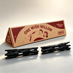 May include: A triangular cardboard box labeled ONE WISH WILLOW with vintage-style illustrations of two people and text reading AMAZE YOUR FRIENDS and You only get ONE WISH. Two black, glossy, cylindrical objects with irregular cutouts lie in front.