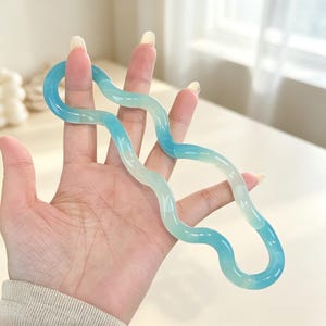 Puede incluir: Un accesorio para el cabello ondulado y translúcido que presenta un degradado de tonos azul y verde claro. El objeto tiene un acabado liso y brillante y una forma serpentina continua que se curva en un diseño fluido y alargado.