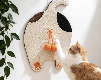 May include: A wall-mounted cat scratching pad shaped like the rear of a cat, crafted from cream-colored rope with brown and black patches. It features two orange yarn balls hanging from a string and orange paw pad details at the bottom of the legs.