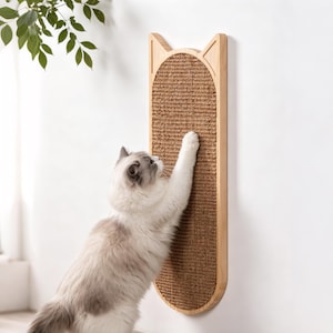 May include: A wall-mounted cat scratching post featuring a light wood frame shaped like cat ears with a textured brown sisal fiber scratching surface. A fluffy white and grey cat is shown reaching up to scratch the vertical panel.