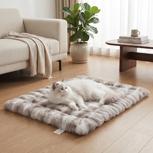 May include: A rectangular pet mat featuring a plush, faux fur texture with a marbled pattern of white, cream, and soft brown tones. A long-haired white and grey cat with blue eyes is resting on the center of the mat, which has a small white tag.