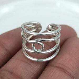 Può includere: Un anello in metallo color argento caratterizzato da un design a fasce multiple con quattro anelli paralleli simili a fili metallici che si sovrappongono e si collegano al centro. L'anello ha una struttura aperta e regolabile.