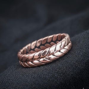 May include: A copper-toned metal ring featuring a continuous, raised braided or wheat-like pattern that wraps around the entire circumference of the band. The surface of the ring has a polished, reflective finish with dark recessed areas between the links.