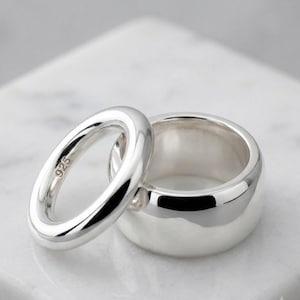 Op de afbeelding: Twee gepolijste sterling zilveren ringen met een glad, afgerond oppervlak. De kleinere ring staat rechtop en heeft een duidelijk 925-keurmerk aan de binnenkant, terwijl de bredere, dikkere ring erachter ligt in een bijpassende zilveren afwerking.