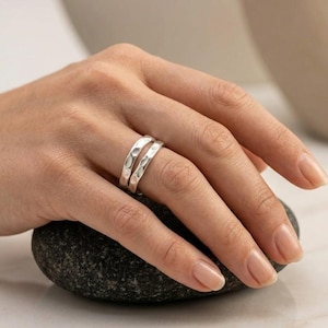 May include: A set of two silver rings with a hammered, textured finish. The rings are worn together on a finger, showcasing a reflective metallic surface with irregular indentations across the bands, creating a distinct, light-catching appearance.