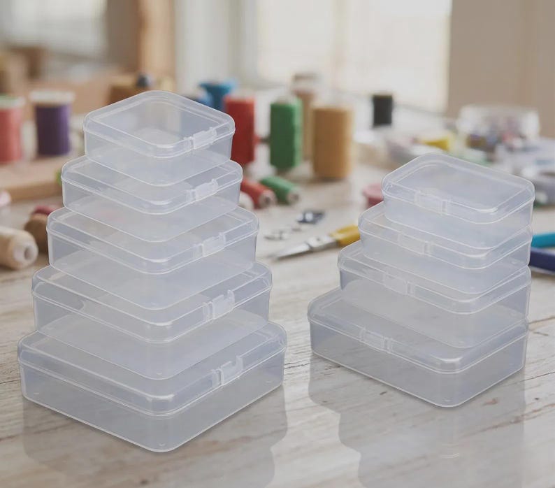 May include: A collection of clear, rectangular plastic storage boxes with hinged lids and snap closures. The boxes are arranged in two separate stacks of varying sizes, featuring a smooth, semi-transparent finish and a simple, functional design.