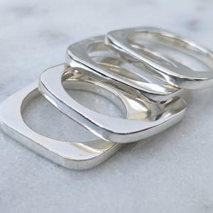 Peut inclure: Un ensemble de quatre bagues en métal argenté poli présentant un design de bande de forme carrée. Les bagues sont empilées en cascade, mettant en valeur une surface métallique lisse et réfléchissante ainsi qu'une épaisseur uniforme.